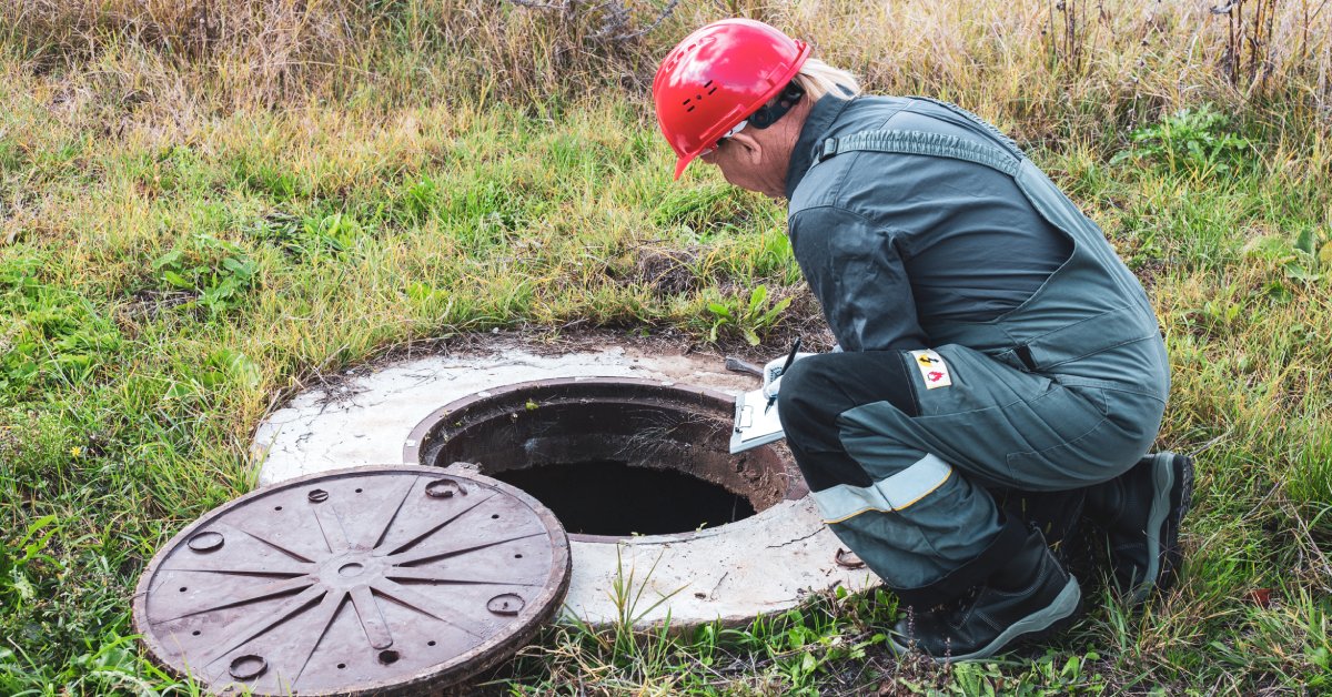 The rear view of a worker kneeling near an open manhole and writing on a clipboard. Grass surrounds the manhole.
